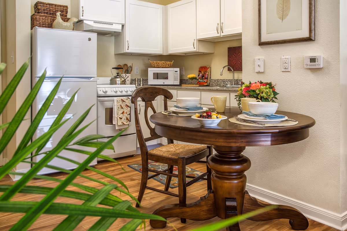 A cozy kitchen and dining area featuring a round wooden table set for two with bowls, plates, mugs, and a bowl of fruit. The kitchen has white cabinets, a white refrigerator, a stove with a kettle, a microwave, and a granite countertop. A green plant is visible in the foreground, and a framed leaf artwork hangs on the wall.