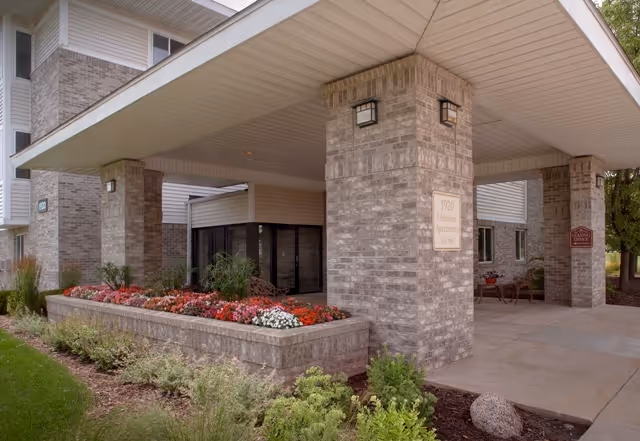 Entrance area of Glenwood Senior Apartments featuring a covered walkway supported by brick pillars, a flower bed with colorful flowers, and surrounding greenery.