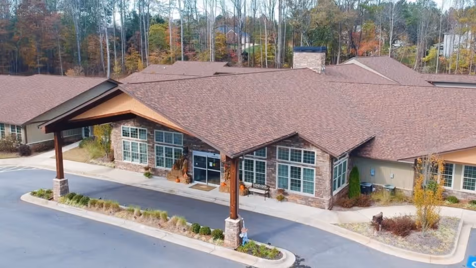Exterior view of The Retreat at Canton facility showing a single-story building with a large covered entrance supported by wooden beams. The building has multiple large windows and a stone facade. Surrounding the building are landscaped areas with shrubs and trees, and a paved driveway in front.