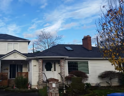 Front exterior view of a single-story house with white siding, a black roof with a skylight, a brick chimney, and a small porch with steps and railings. There are trimmed bushes and a tree with autumn leaves in the front yard under a partly cloudy blue sky.