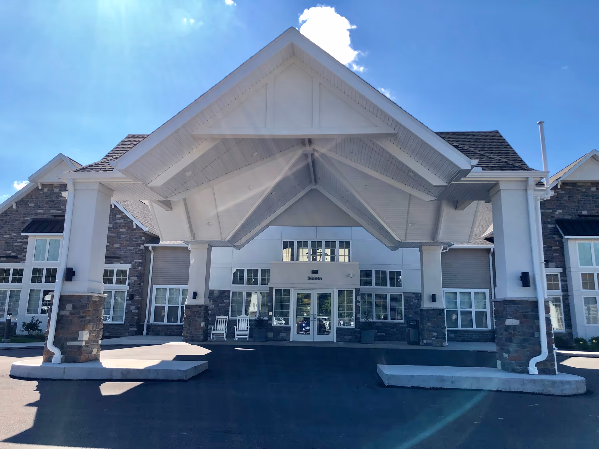 Front exterior view of a senior living facility building with a large covered entrance supported by stone and white pillars, multiple windows, and a clear blue sky above.