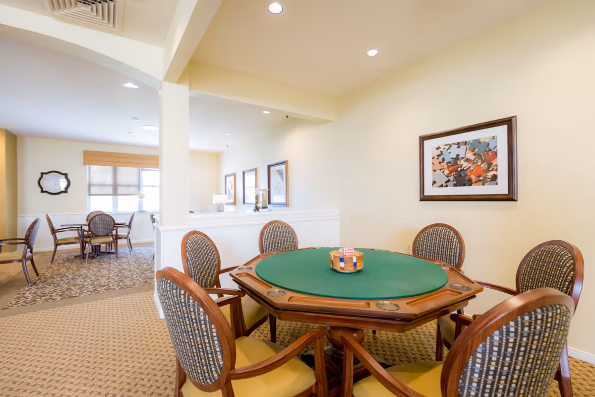 A well-lit room with a green felt octagonal card table surrounded by six wooden chairs with patterned upholstery. In the background, there are additional tables and chairs near windows with blinds, and framed artwork on the walls. The room has beige walls and carpeted floors with a patterned design.