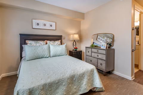 A cozy bedroom with a neatly made bed featuring a light green quilt and matching pillows. There is a dark wooden headboard, a nightstand with a lamp and a small flower arrangement, and a dresser with a mirror, a framed photo, and decorative plants. The room has beige walls and carpeted flooring. A doorway leads to a bathroom area.