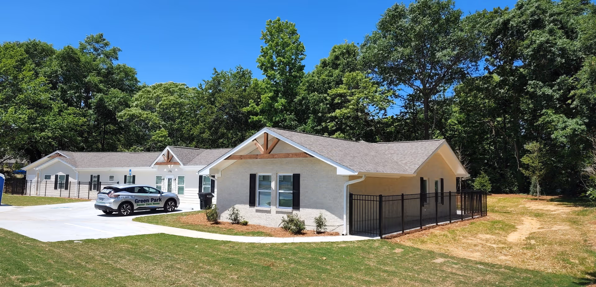 Exterior view of a single-story senior care home building with light-colored walls and a gray roof, surrounded by green grass and trees under a clear blue sky. A car with 'Green Park Senior Care Home' branding is parked on the driveway.