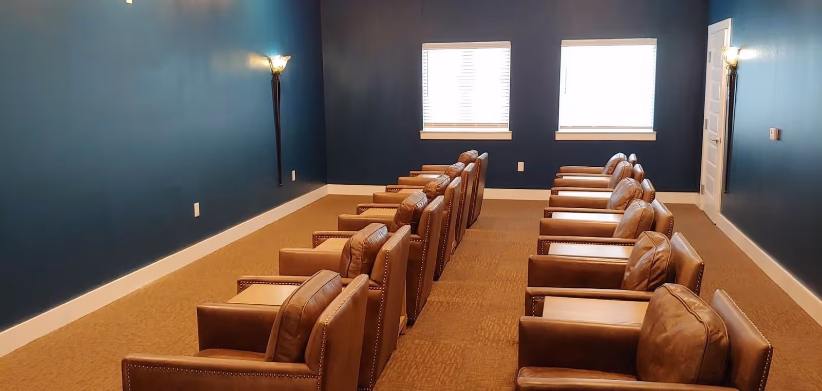 Room with two rows of brown leather armchairs facing forward, blue walls, beige carpet, two windows with blinds, and wall-mounted light fixtures.