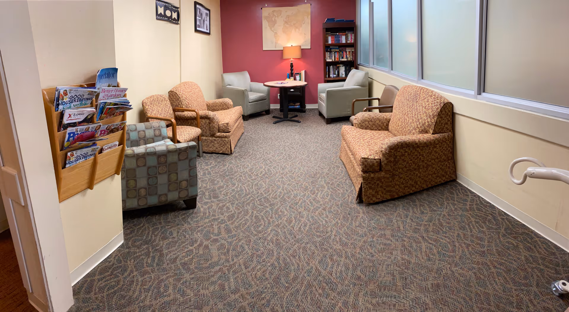 Carpeted common lounge with several armchairs, a small table with a lamp, a bookshelf and a wall-mounted magazine rack.
