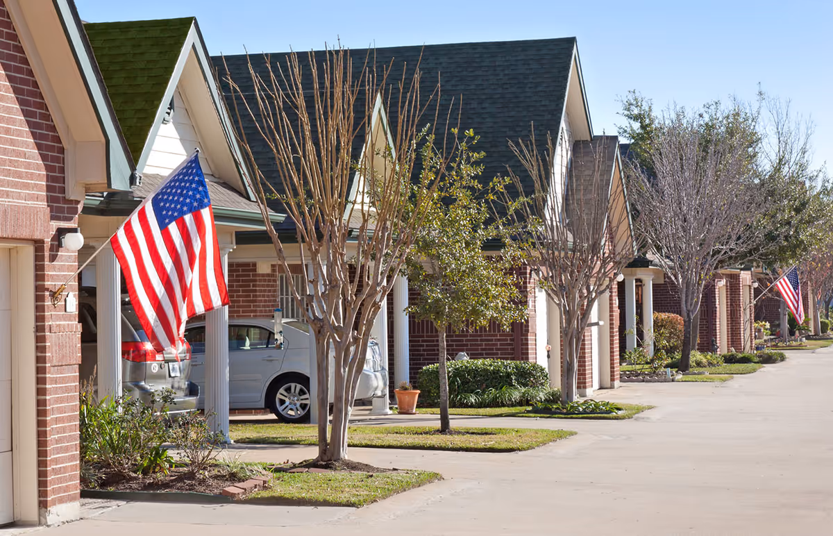 Row of single-story brick residential buildings with green and dark gray roofs, each with a driveway and an American flag mounted near the entrance. Leafless trees and some greenery line the sidewalk in front of the homes under a clear blue sky.