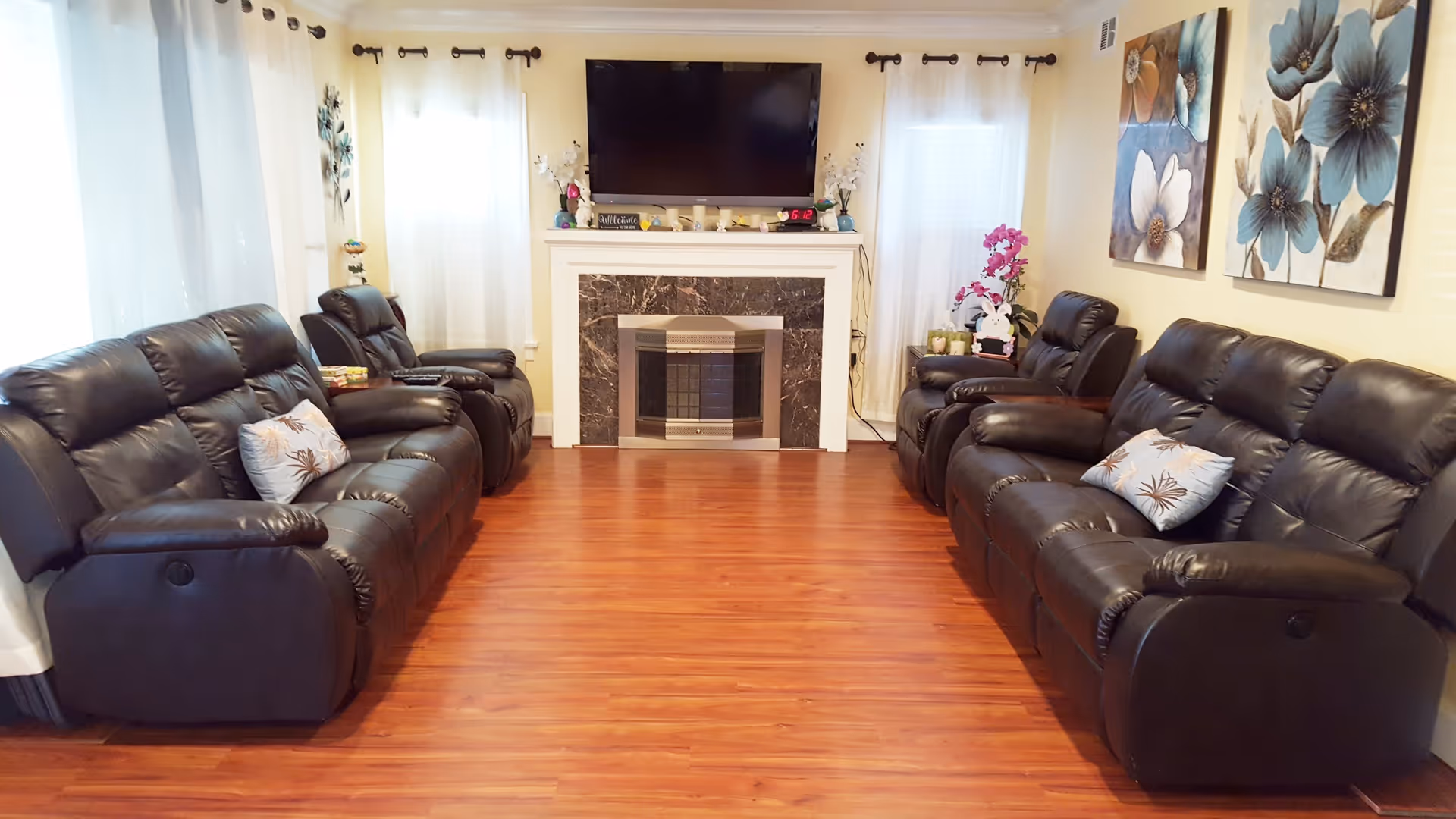 A cozy living room with two black leather sofas and two matching recliners facing each other across a wooden floor. At the far end, there is a fireplace with a dark marble surround and a flat-screen TV mounted above it. The room is decorated with floral paintings on the right wall, white curtains on the windows, and various small decorative items on the fireplace mantel and side tables.