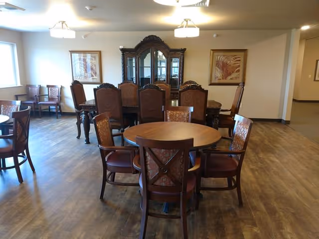 A dining room with multiple wooden tables and chairs arranged on a wood floor. The room has beige walls with two framed artworks and a large dark wooden china cabinet against the far wall. There are windows letting in natural light and ceiling lights providing additional illumination.