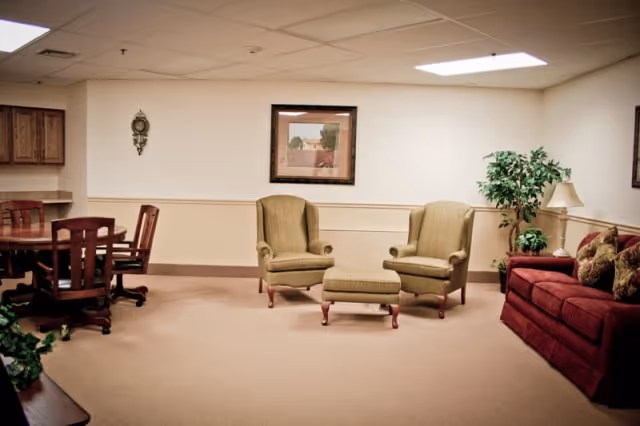 A cozy living room area in a retirement community featuring two beige armchairs with a matching ottoman, a red sofa with patterned cushions, a wooden dining table with chairs, a potted plant, a table lamp, and framed artwork on the wall.