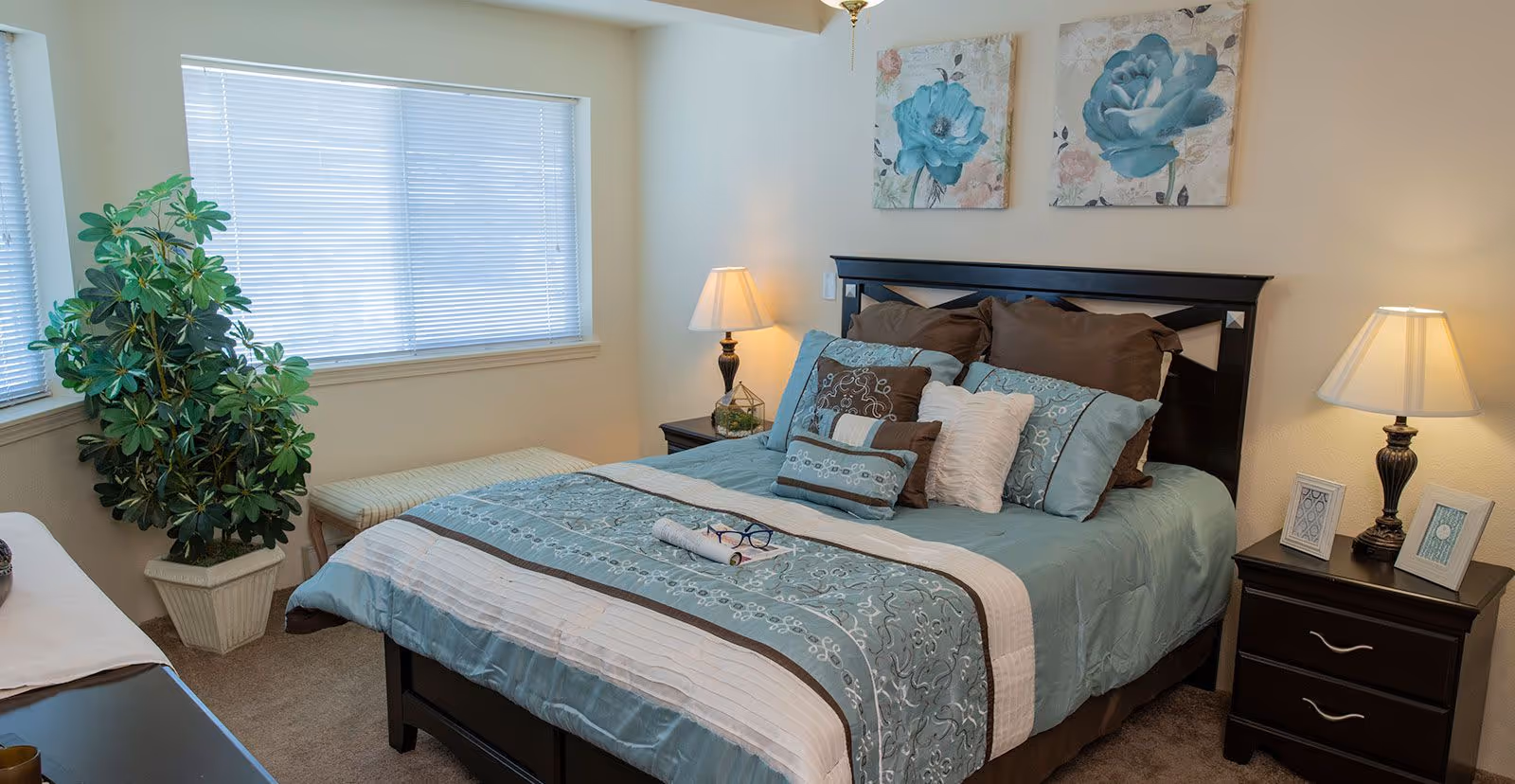 A cozy bedroom with a neatly made bed featuring blue and brown bedding and multiple pillows. Two framed floral paintings hang above the headboard. There are two bedside tables with lamps and picture frames. A large window with blinds lets in natural light, and a green potted plant is placed near the window. A bench is positioned under the window.