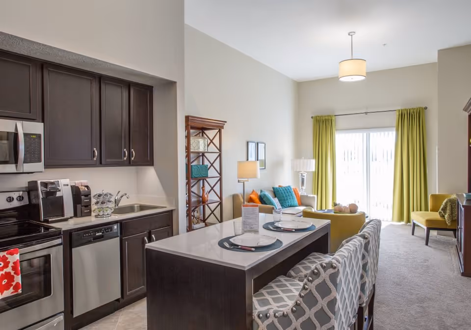 A modern open-concept living space featuring a kitchen with dark wood cabinets, stainless steel appliances, and a sink. Adjacent to the kitchen is a dining area with a white countertop island and two patterned chairs. The living room area includes a beige sofa with colorful cushions, a yellow armchair, a wooden shelving unit, and large windows with green curtains allowing natural light to fill the room.