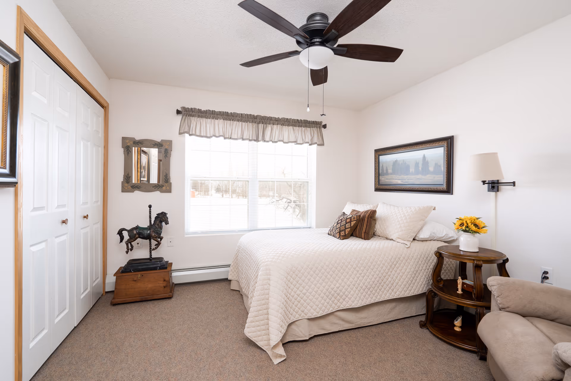 Bright, neatly furnished bedroom with a bed, ceiling fan, window, bedside table with flowers, and a closet.
