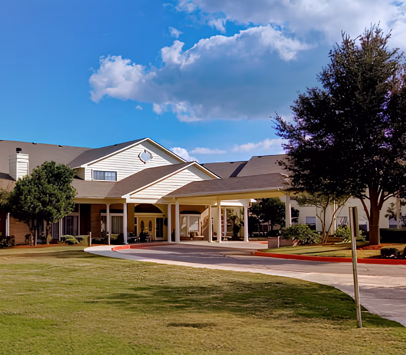 Exterior view of a senior living facility building with a covered entrance driveway, surrounded by green grass and trees under a blue sky with some clouds.
