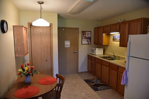 Small assisted-living kitchen and dining area with wooden cabinets, sink and microwave on the right, a round table with placemats and flowers on the left, and closed wooden doors in the center.