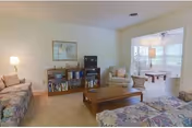 A cozy living room with floral patterned sofas, a wooden coffee table, a bookshelf with books and decorative items, a small TV, and a lamp. Adjacent to the living room is a bright room with a pool table and large windows letting in natural light.