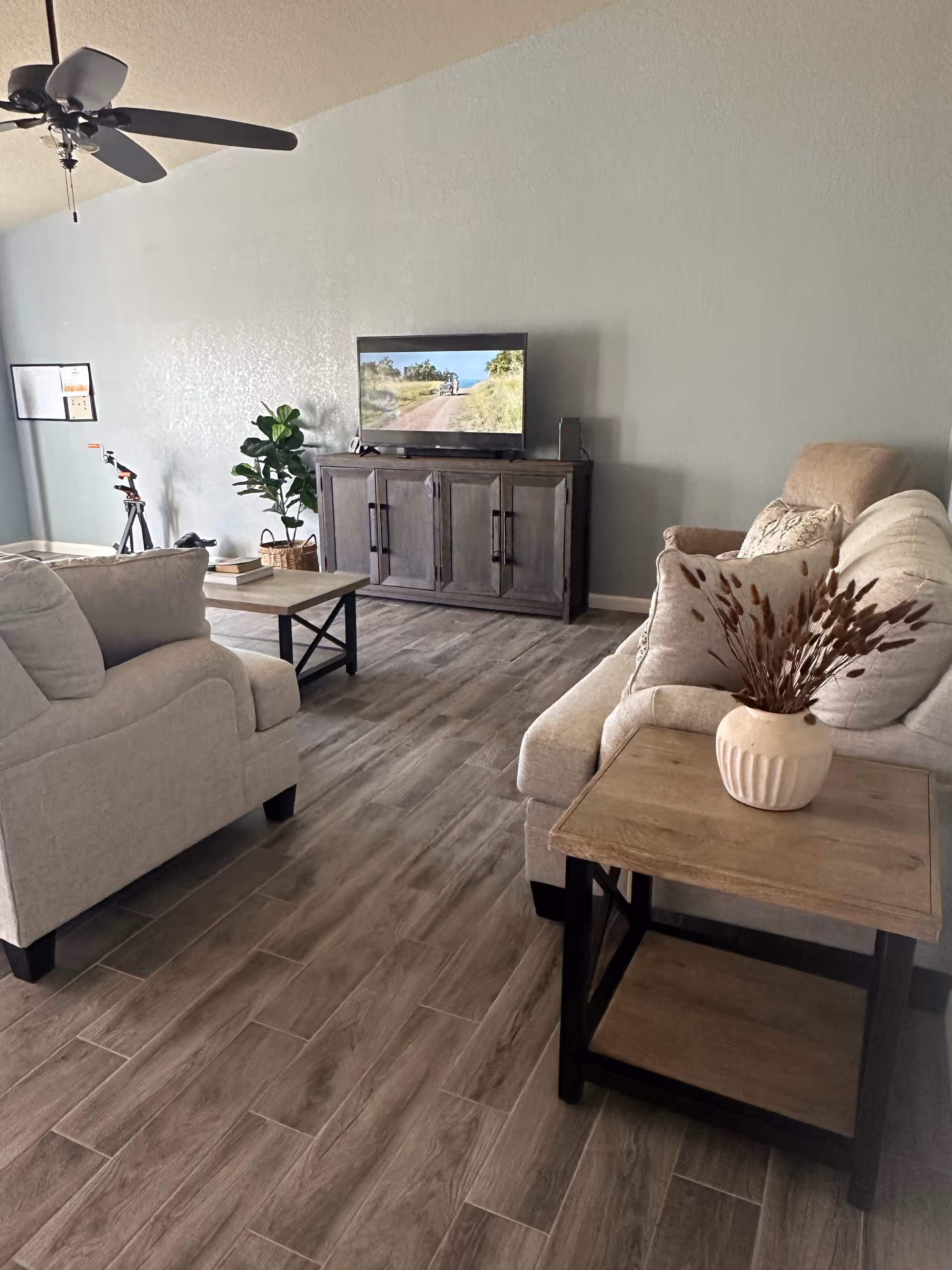 A cozy living room with light-colored sofas and armchairs arranged around a wooden coffee table. A wooden TV stand holds a flat-screen television displaying a nature scene. There is a ceiling fan above, a potted plant in the corner, and a side table with a decorative vase containing dried plants. The floor is covered with wood-like tiles, and the walls are painted a soft gray.