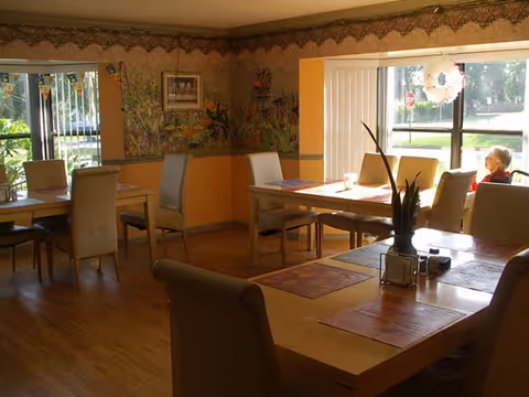 A dining room in a senior care facility with several tables and chairs arranged neatly. The walls are decorated with floral murals and a border near the ceiling. Large windows allow natural light to fill the room, and an elderly person is seated near one of the windows.