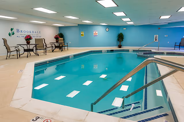Indoor community swimming pool with metal handrails and seating tables along a light blue wall.
