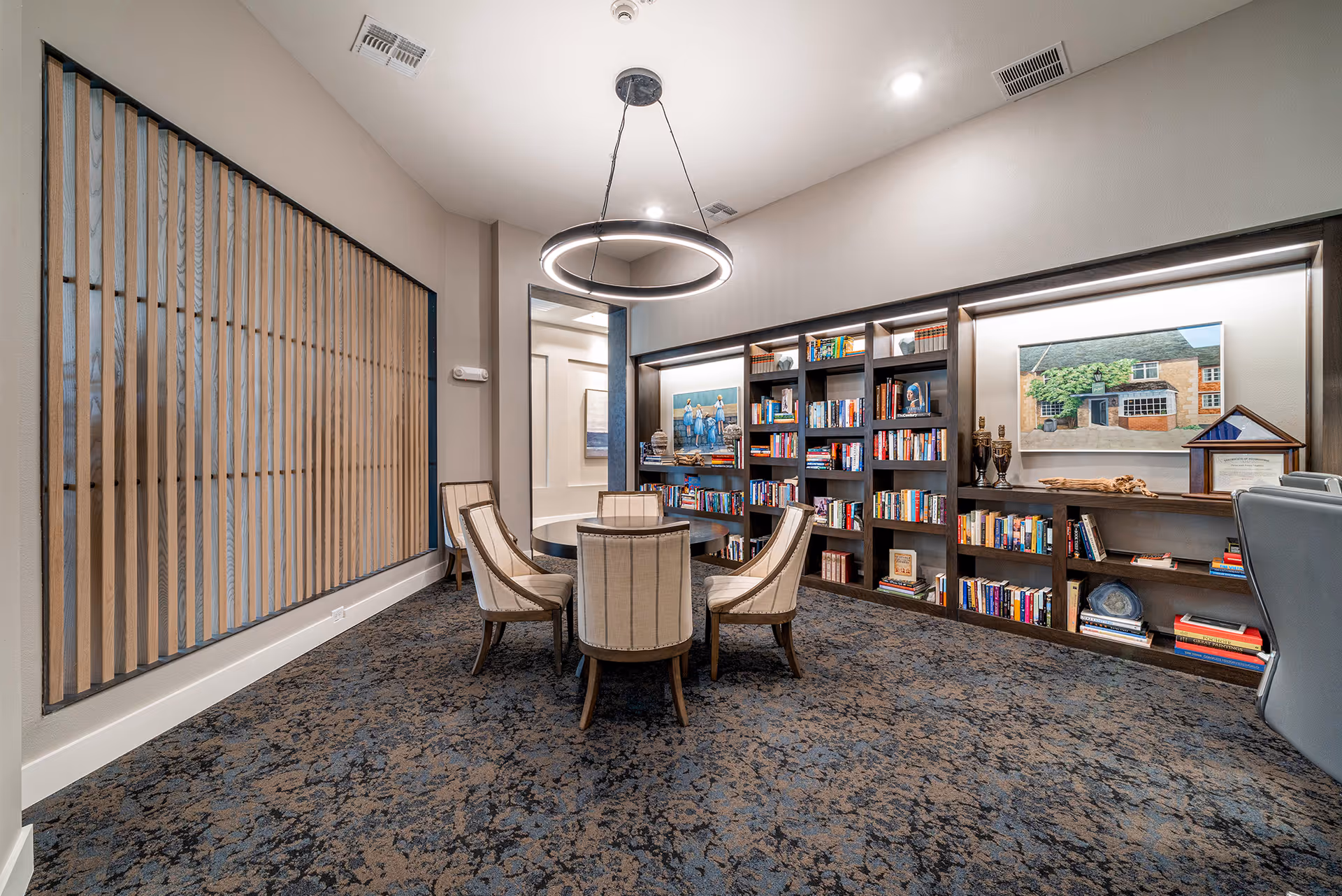 A cozy communal reading room with a round table and upholstered chairs in front of built-in bookshelves and framed artwork.