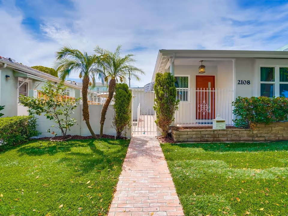 Front exterior view of a single-story residential building with a red door, white fence, green lawn, palm trees, and shrubs under a partly cloudy sky.