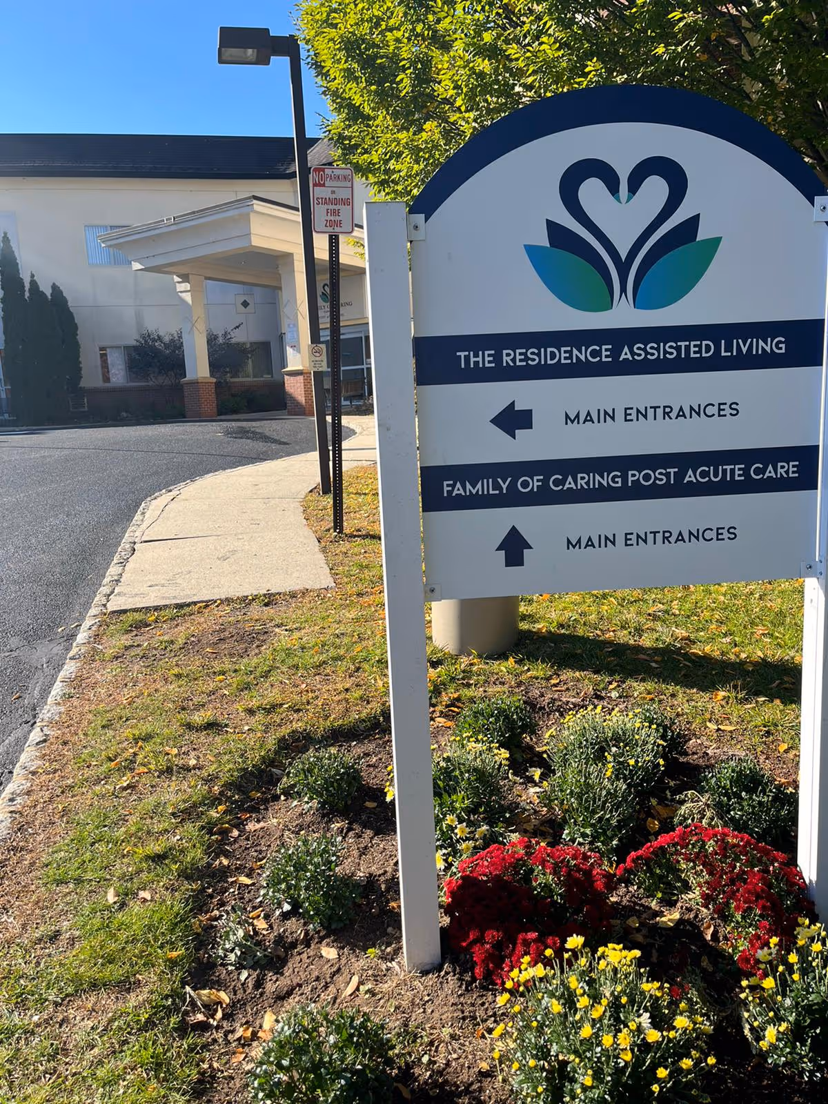 Outdoor view of the entrance area of Family of Caring at Park Ridge facility with a directional sign indicating main entrances for The Residence Assisted Living and Family of Caring Post Acute Care. The sign is surrounded by flower beds with red and yellow flowers, and a building is visible in the background.