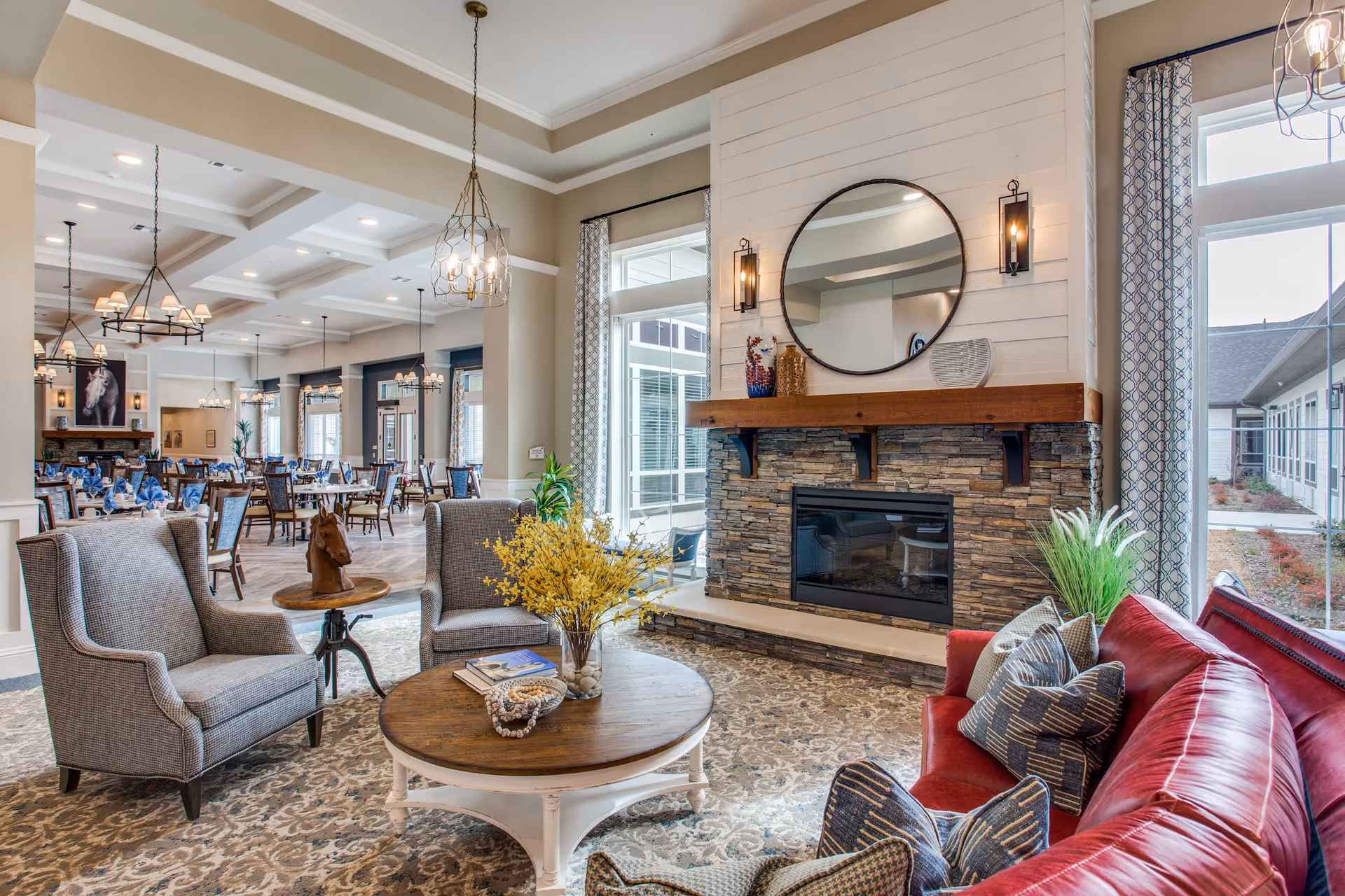 A cozy senior living common area featuring a stone fireplace with a round mirror above it, flanked by two wall sconces. In front of the fireplace is a round wooden coffee table with a vase of yellow flowers and decorative items. Surrounding the table are two gray armchairs and a red leather sofa with patterned pillows. Large windows with patterned curtains allow natural light to fill the room. In the background, there is a dining area with multiple tables and chairs, elegant chandeliers, and a painting of a horse above a fireplace.