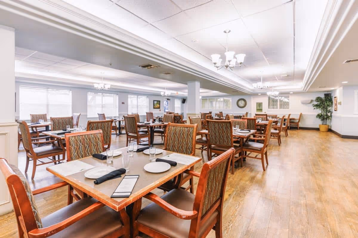 A spacious dining room with multiple wooden tables and chairs arranged neatly. Each table is set with plates, glasses, napkins, and menus. The room has large windows allowing natural light to fill the space, light-colored walls, and ceiling lights with chandeliers. There is a clock on the far wall and a potted plant in the corner.