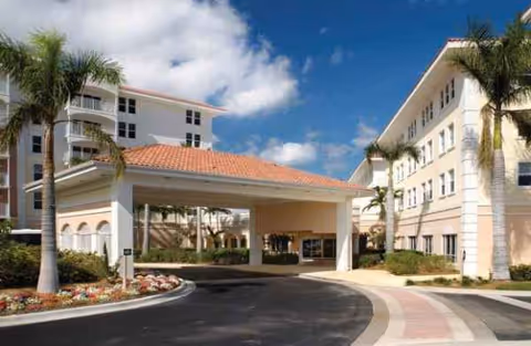 Two-story retirement community with a covered entrance and palm trees along a circular driveway.