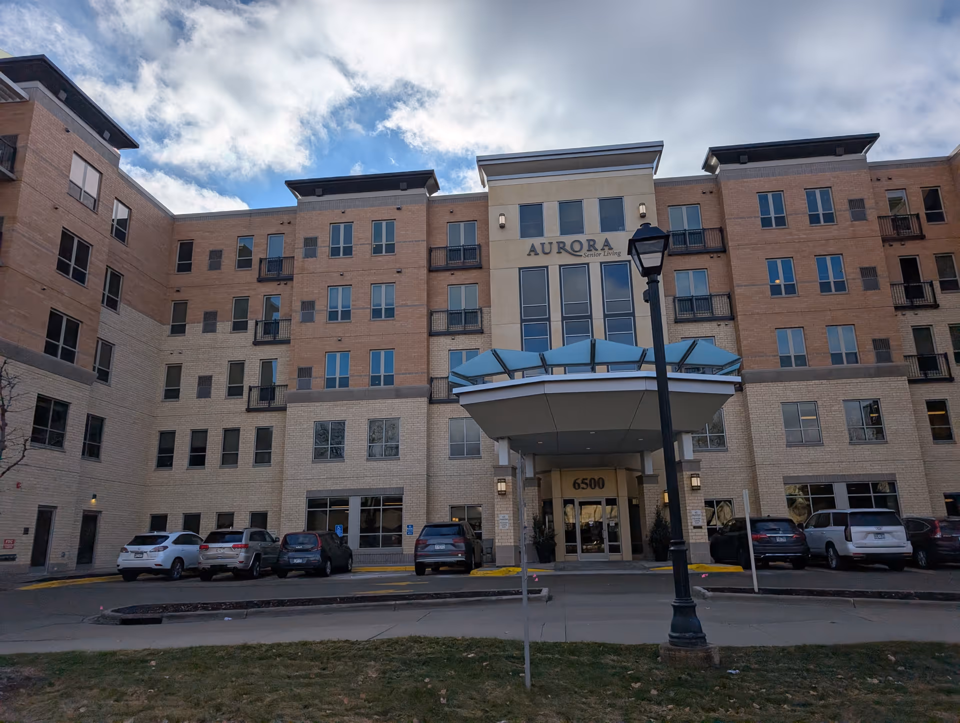 Front exterior view of a multi-story senior living facility named Aurora on France with a covered entrance, several parked cars, and a street lamp in front under a partly cloudy sky.