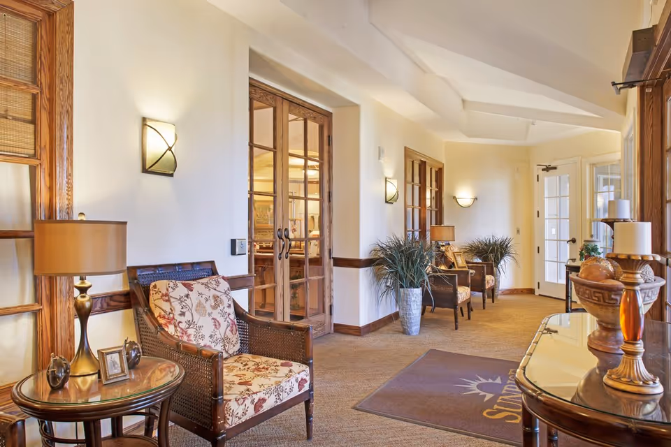 A well-lit hallway in a senior living facility with wooden framed glass doors, floral cushioned chairs, side tables with lamps, potted plants, and decorative items on a console table. The floor has a carpet with a mat that reads 'Sunrise'.
