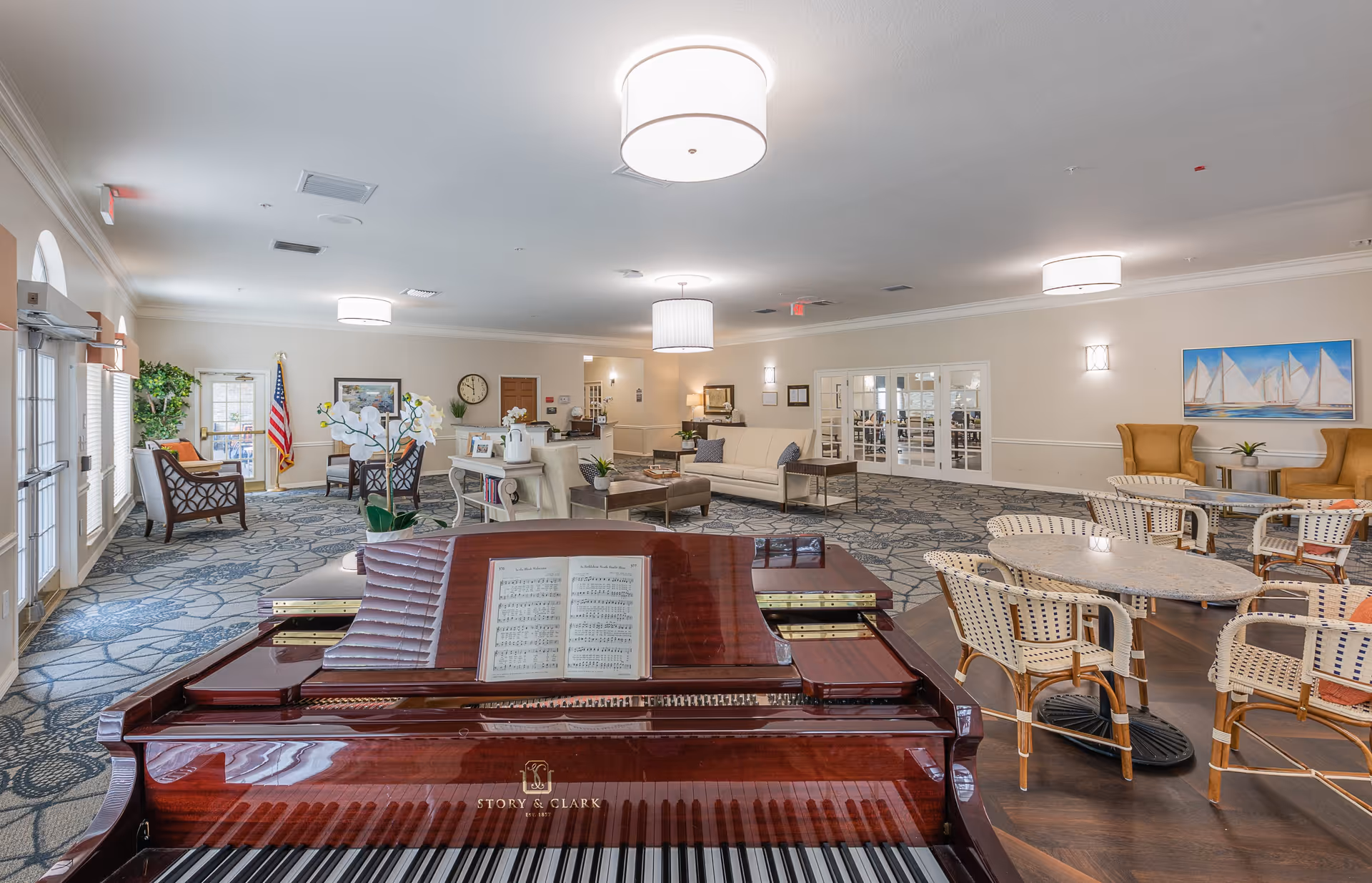 Interior view of a spacious senior living facility common area featuring a polished Story & Clark grand piano in the foreground with sheet music on the stand. The room includes multiple seating arrangements with chairs and sofas, round tables with wicker chairs, a carpeted floor with a floral pattern, and soft overhead lighting. There is an American flag near the entrance door, framed artwork on the walls, and large windows letting in natural light.