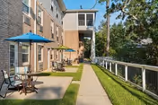 Outdoor patio area at The Camellia at Gulf Breeze featuring a paved walkway, green grass, several small tables with chairs and blue umbrellas, adjacent to a multi-story brick building with screened balconies and surrounded by trees.