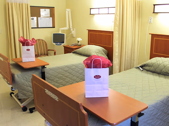 Two hospital-style beds in a softly lit shared patient room with overbed tables, bedside furniture, and gift bags on the trays.