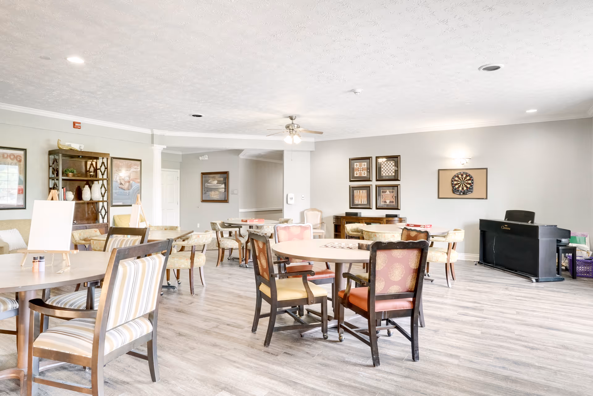 A bright and spacious common area with multiple round tables and chairs arranged for group activities. The room features light-colored walls, wood flooring, a ceiling fan, framed artwork on the walls, a dartboard, and a black piano in the corner.