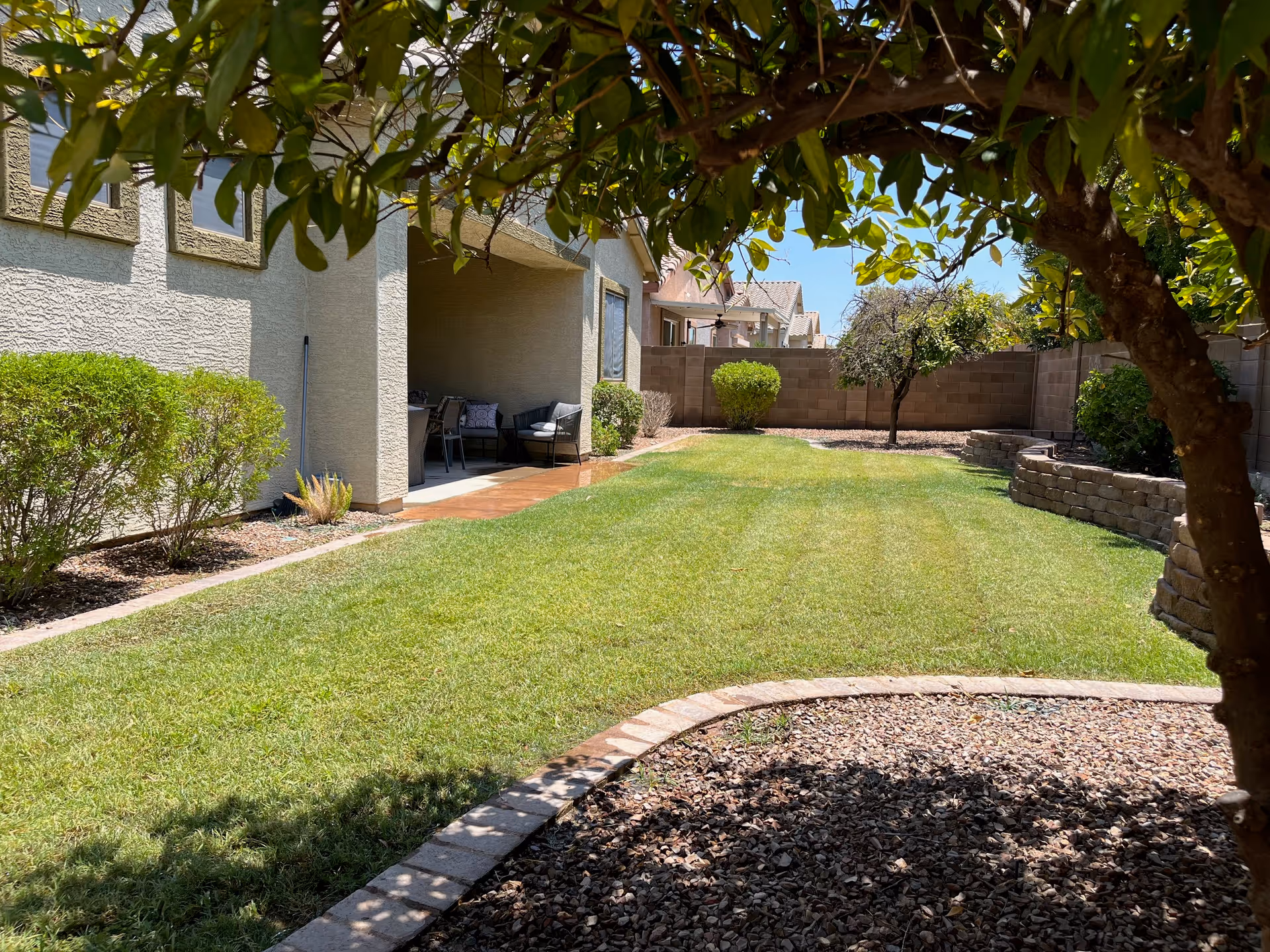 A sunny backyard with a well-maintained green lawn, bordered by bushes and a tree casting shade in the foreground. There is a covered patio area attached to a beige stucco house with outdoor seating. A stone retaining wall lines one side of the yard, and a block wall encloses the space.