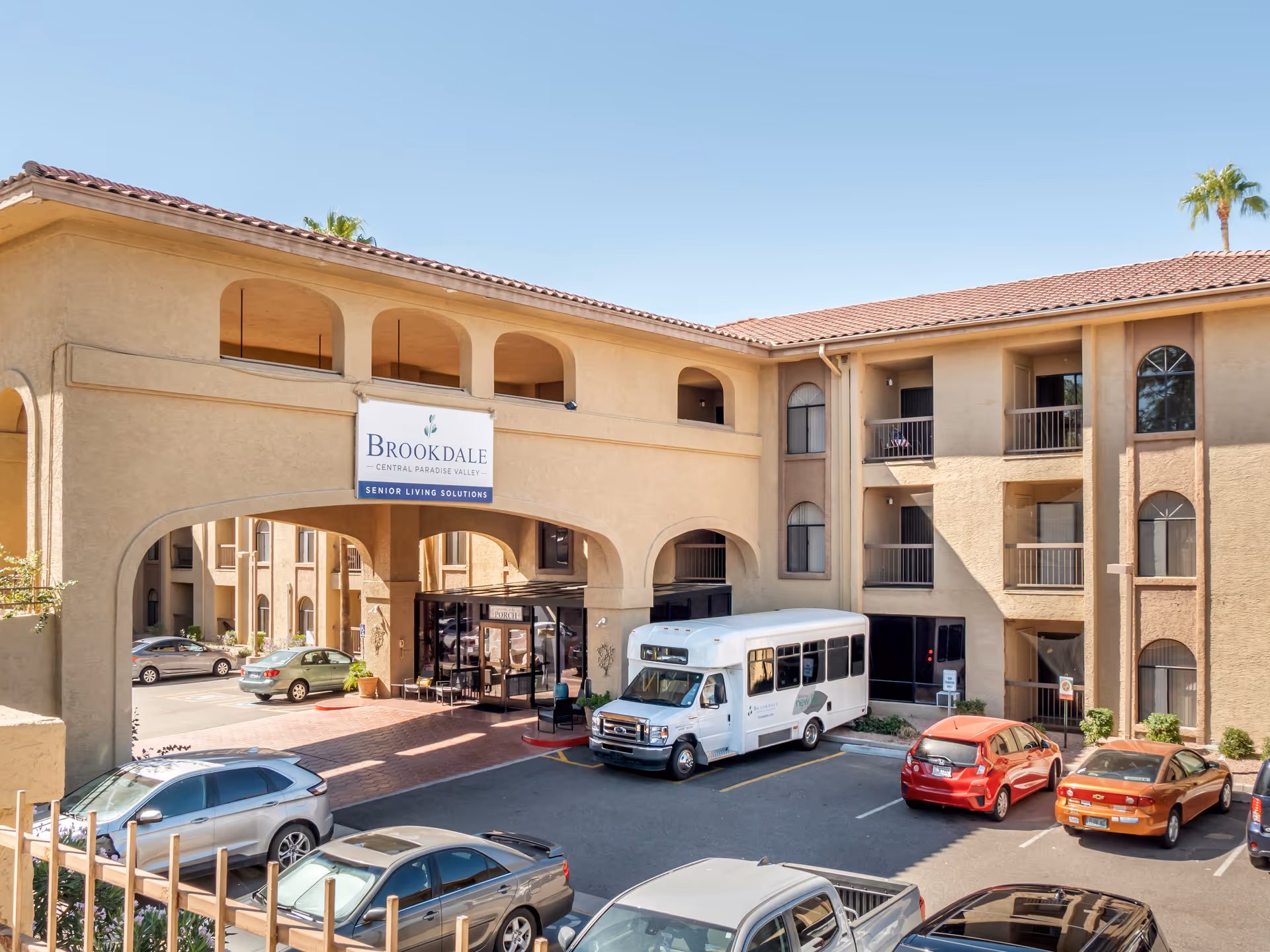 Exterior view of Brookdale Central Paradise Valley senior living facility showing a beige stucco building with arched openings and a tiled roof. Several cars and a white shuttle bus are parked in front of the entrance under a covered archway. The sky is clear and blue.