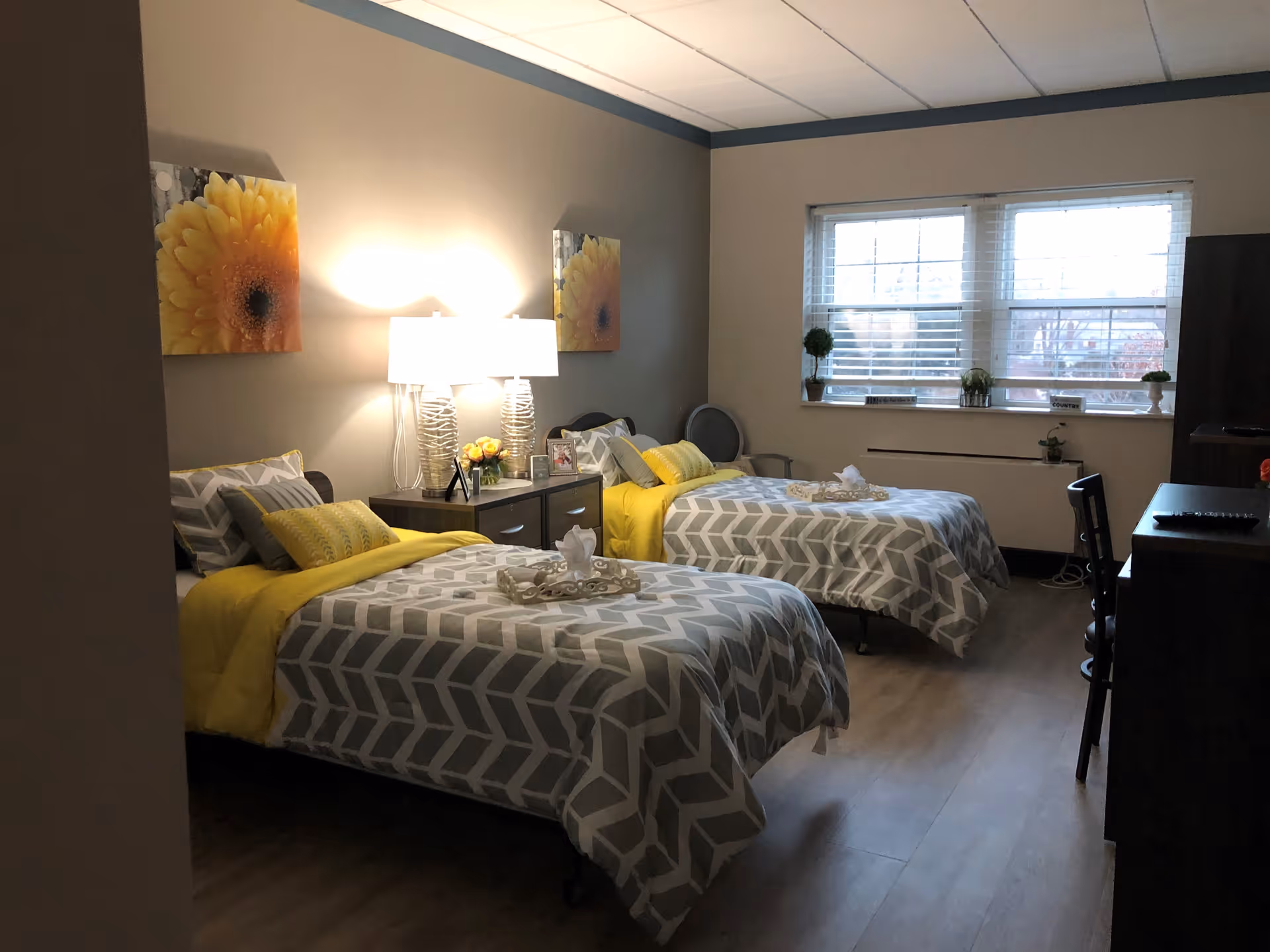 A bedroom with two single beds, each made with gray and white patterned bedding and yellow blankets. Between the beds is a nightstand with two lamps and decorative items. The room has a window with blinds, a chair in the corner, and a dresser with a chair on the right side. The walls are decorated with yellow flower artwork.