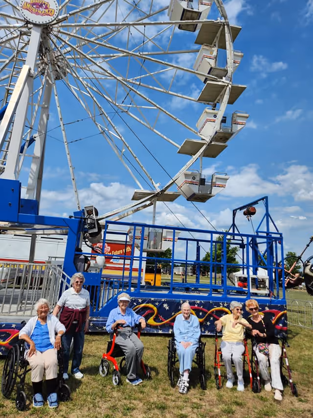 A group of six elderly women, some seated in wheelchairs and others using walkers, posing and smiling in front of a large Ferris wheel at an outdoor amusement area on a sunny day with blue skies and scattered clouds.