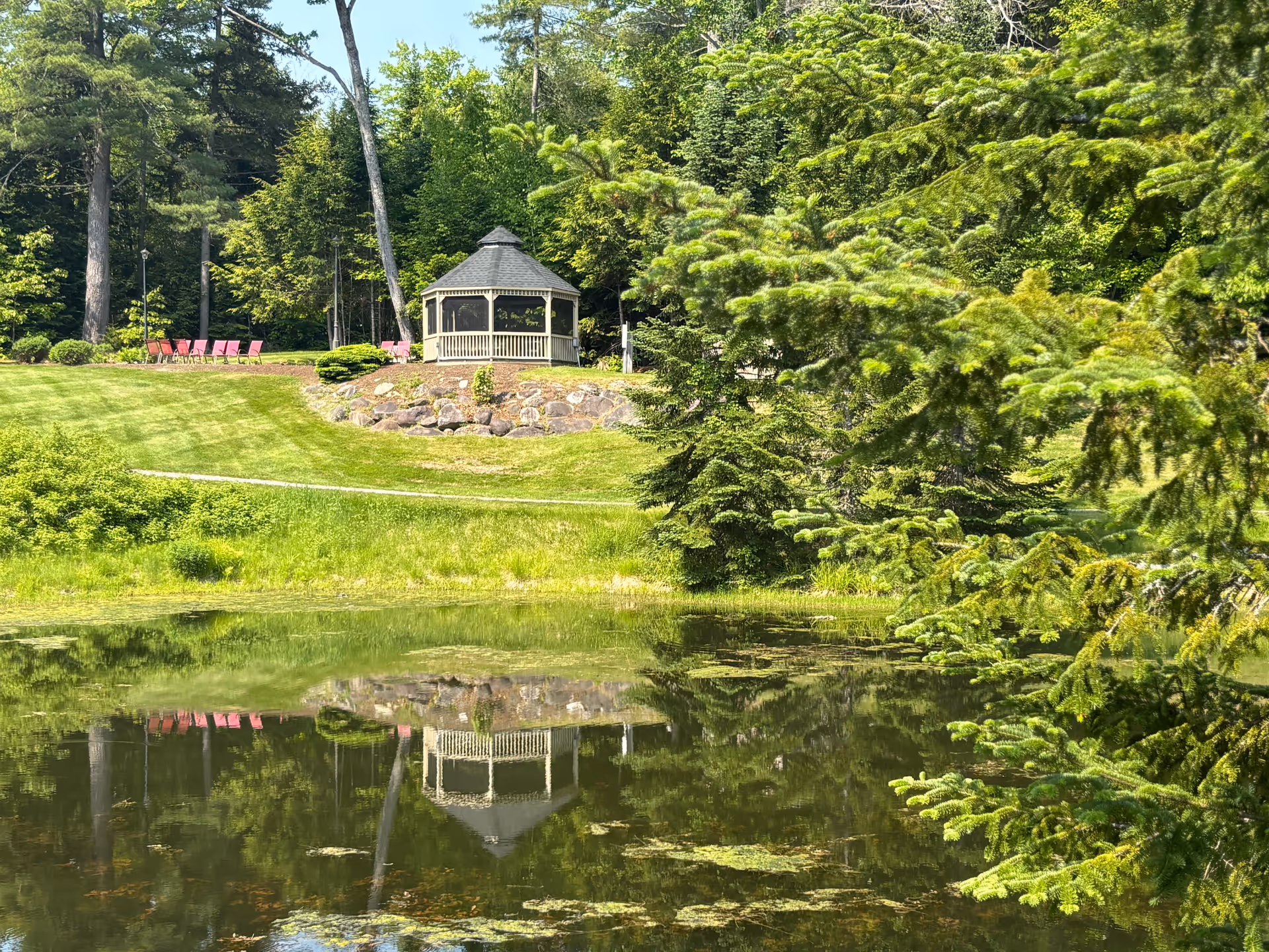 A serene outdoor scene featuring a small pond reflecting a white gazebo surrounded by lush green trees and grass. There are several red chairs arranged on a grassy hill near the gazebo under a clear blue sky.