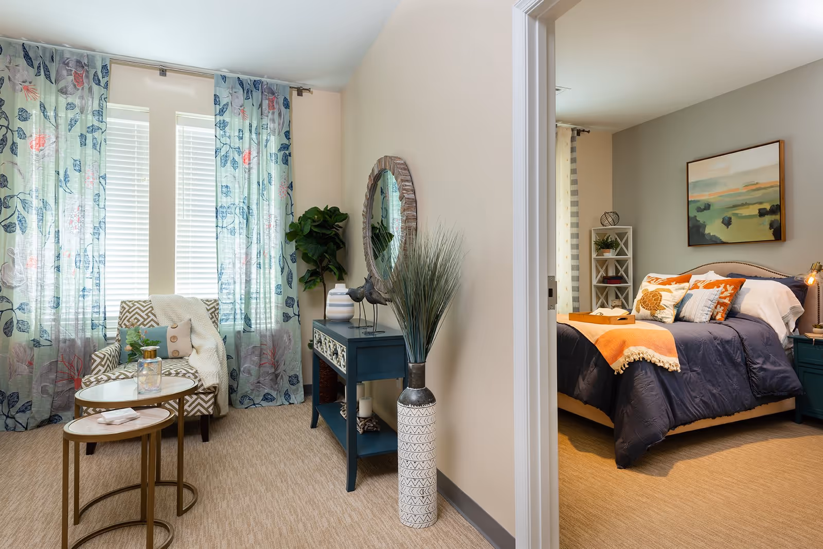 A cozy living area with a patterned armchair, two nested round tables, and sheer floral curtains covering two windows. A blue console table with decorative items and a round mirror hangs on the wall. Adjacent to the living area is a bedroom with a neatly made bed featuring blue and orange bedding, a nightstand, and a framed landscape painting on the wall.