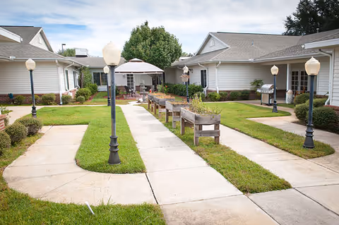 Outdoor courtyard area at a senior living facility with paved walkways, green grass, lamp posts, raised garden beds, patio tables with umbrellas, and single-story buildings surrounding the space.