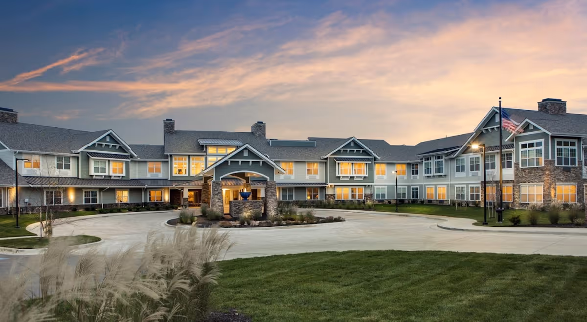 Front exterior of a large two-story senior living facility with a circular driveway, landscaped entrance, and an American flag at sunset.