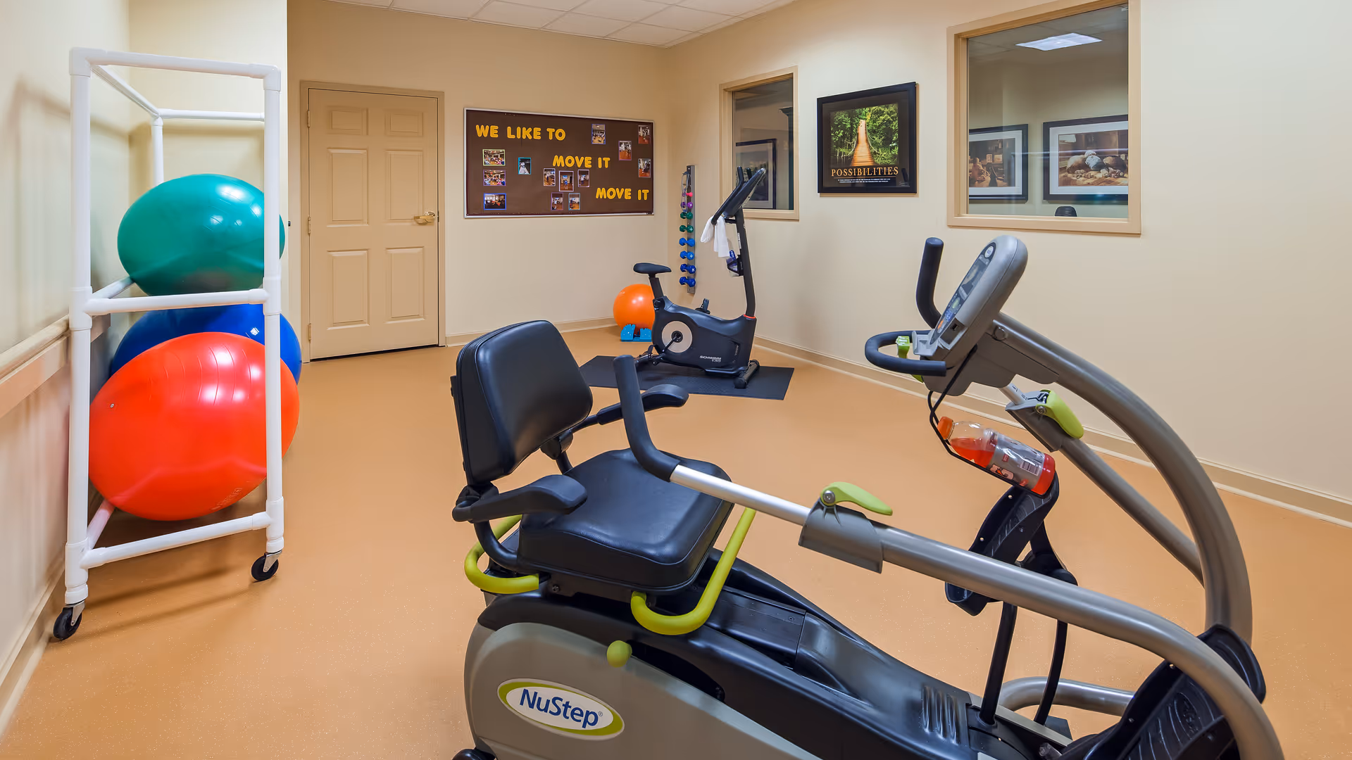 A small exercise room with a recumbent stationary bike in the foreground, another upright stationary bike in the background, a rack holding large exercise balls in red, blue, and green, and a bulletin board on the wall with the phrase 'WE LIKE TO MOVE IT MOVE IT'. The room has beige walls and flooring, with two windows and framed pictures on the wall.