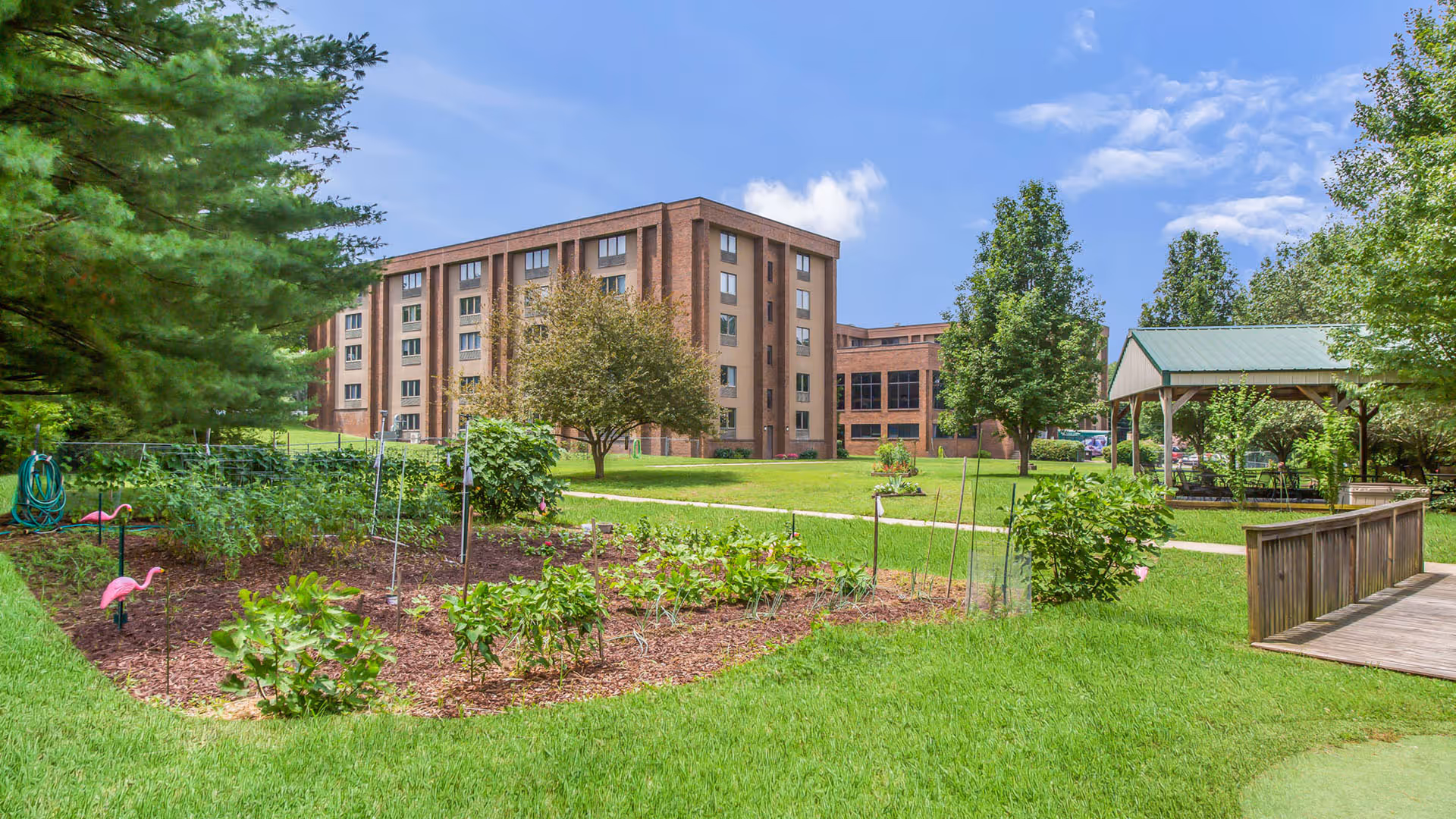 A brick senior living building set behind a grassy courtyard with garden plots and a gazebo under a blue sky.