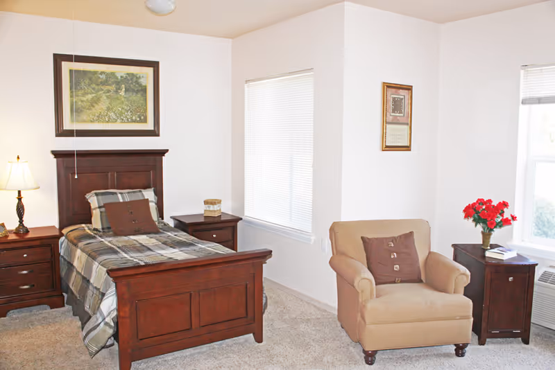A cozy bedroom with a single wooden bed featuring plaid bedding and a brown pillow. There are two wooden nightstands on either side of the bed, one with a lamp and the other with a small decorative item. A beige armchair with a brown pillow is positioned near a window with blinds. A small wooden cabinet with a vase of red flowers and some books is next to the armchair. The walls are light-colored with framed artwork hanging above the bed and near the armchair.