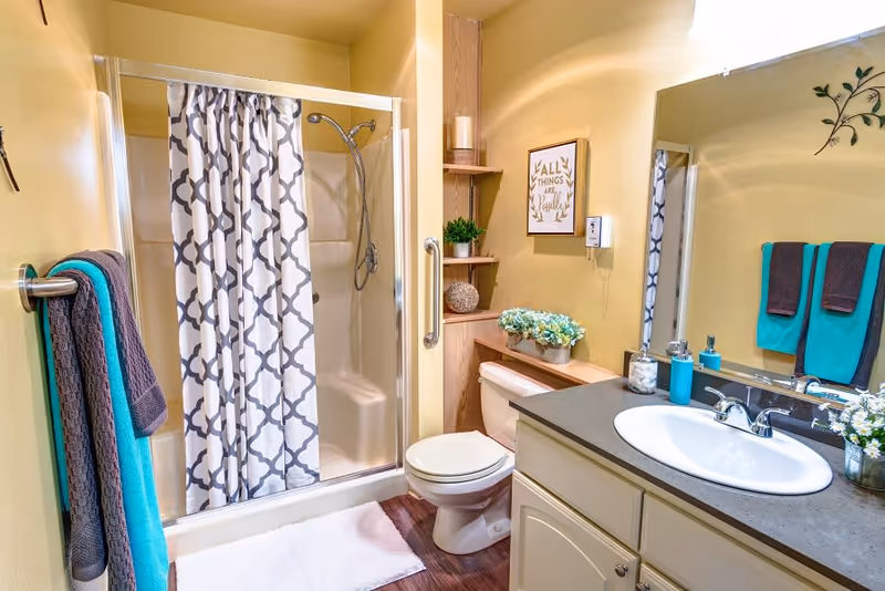 A clean and well-lit bathroom featuring a shower with a patterned curtain, a toilet, and a sink with a countertop. The walls are painted yellow, and there are teal and gray towels hanging on a towel rack. Decorative items include a framed sign, plants, and a candle on wooden shelves next to the toilet.