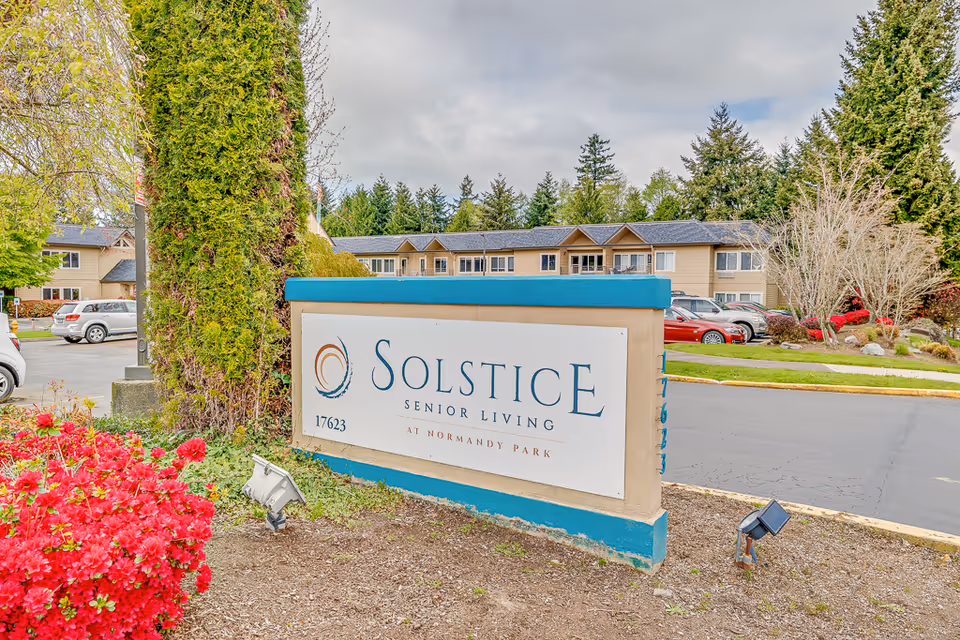 Outdoor view of the entrance sign for Solstice Senior Living at Normandy Park, surrounded by greenery, flowers, and a parking lot with cars in the background.