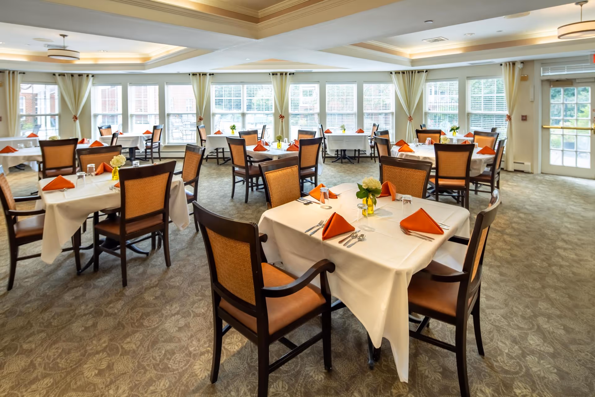 A bright dining room with multiple tables covered in white tablecloths, each set with orange folded napkins, silverware, glasses, and small floral centerpieces. The room has large windows with white curtains allowing natural light to fill the space, and carpeted flooring with a subtle pattern.