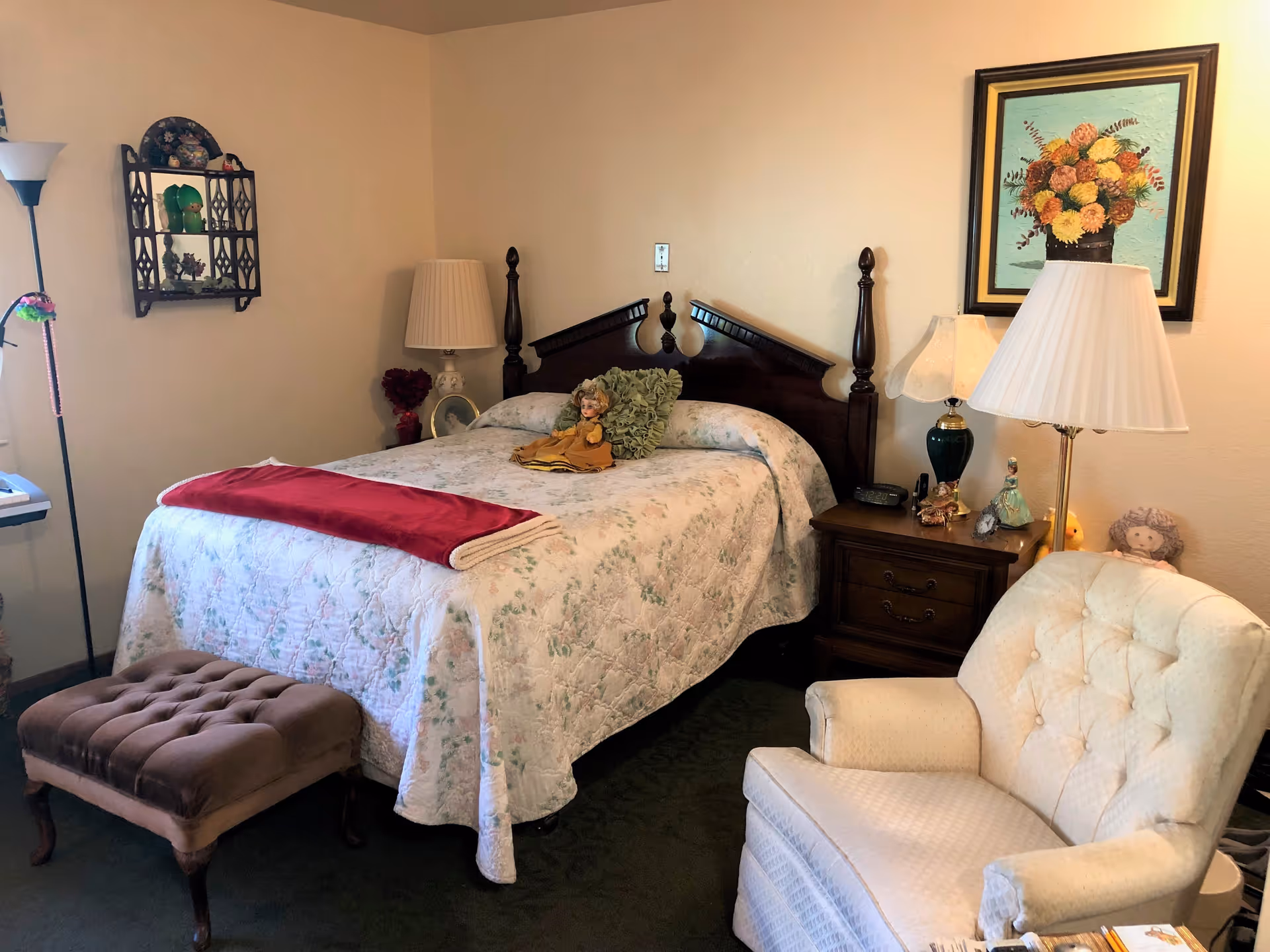 A cozy bedroom with a wooden bed frame, floral quilt, and a red blanket folded at the foot of the bed. There are two bedside tables with lamps, a cushioned armchair, a small ottoman, and decorative items including dolls and a framed floral painting on the wall.